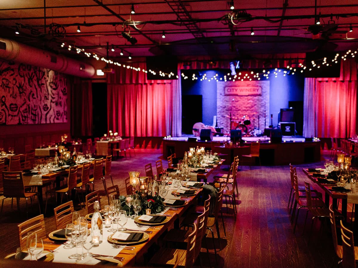 The City Winery Chicago room before a show — long banquet tables set with wine glasses and candles, the lit stage at back framed by plum velvet curtains and the illuminated 'CITY WINERY' sign, exposed-pipe ceiling.