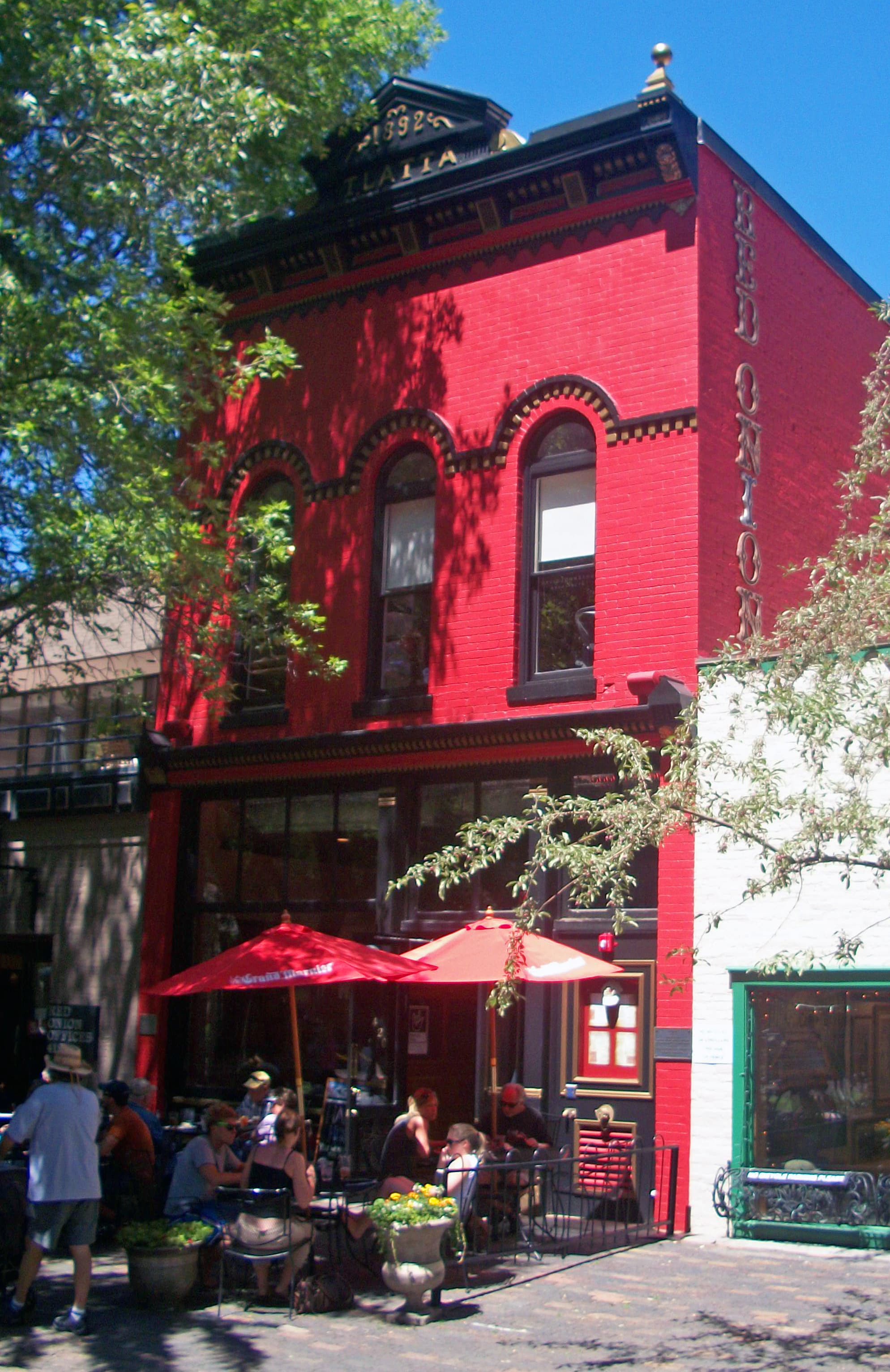 The Red Onion Building at 420 East Cooper Avenue in Aspen — a vermillion-red 19th-century brick storefront with arched second-story windows and the words RED ONION lettered on the cornice, sidewalk cafe with red umbrellas in front under a clear summer sky.